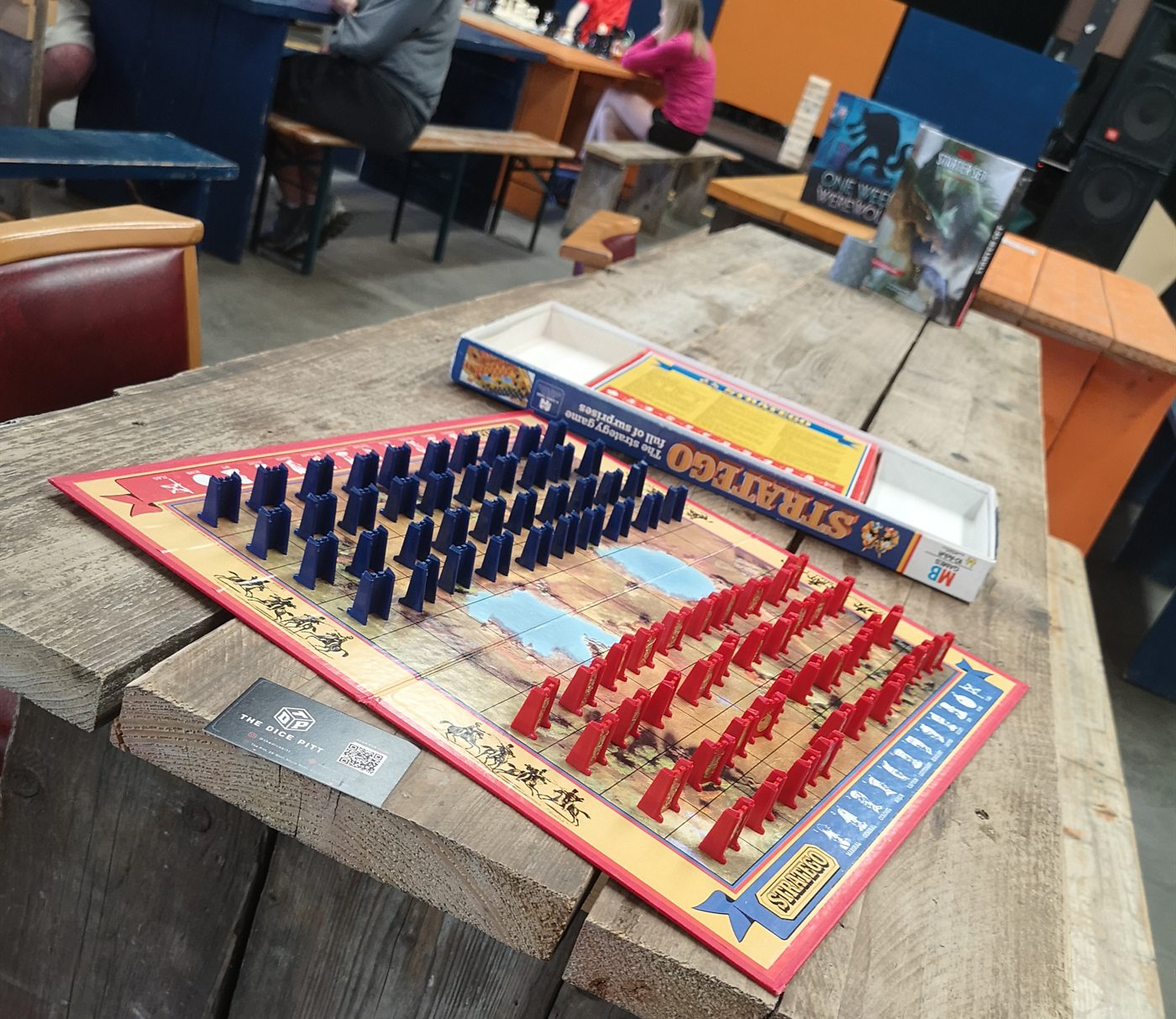 A board game set up on a wooden table at The Dice Pitt with players in the background.