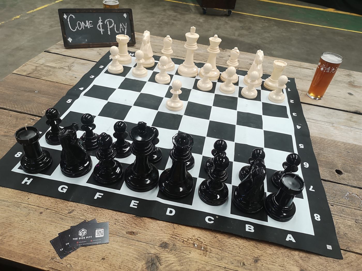 A chess board set up on a wooden table with Dice Pitt cards nearby.